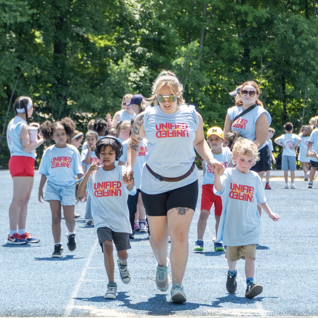 A group of people, including children and adults, stand in line waiting to attempt the long jump. An MPS staff member leads two children wearing matching shirts.