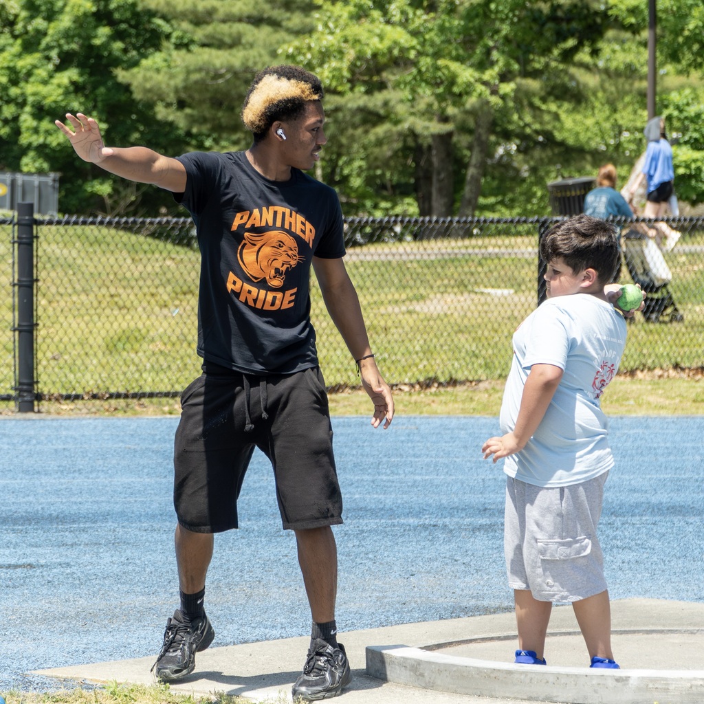 An MHS student wearing all black and a Panther Pride" shirt stands with one arm raised, talking to a child attempting a shot put.