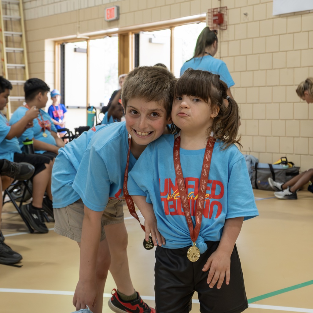 Two children stand in a gymnasium, smiling, with medals around their necks. Behind them, others sit or stand.