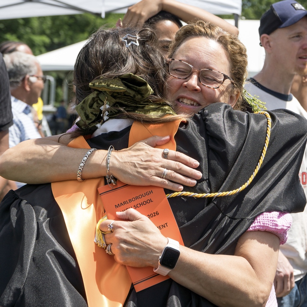 A woman embraces an MHS graduate wearing a graduation gown. Behind them, people stand outside or under a tent.