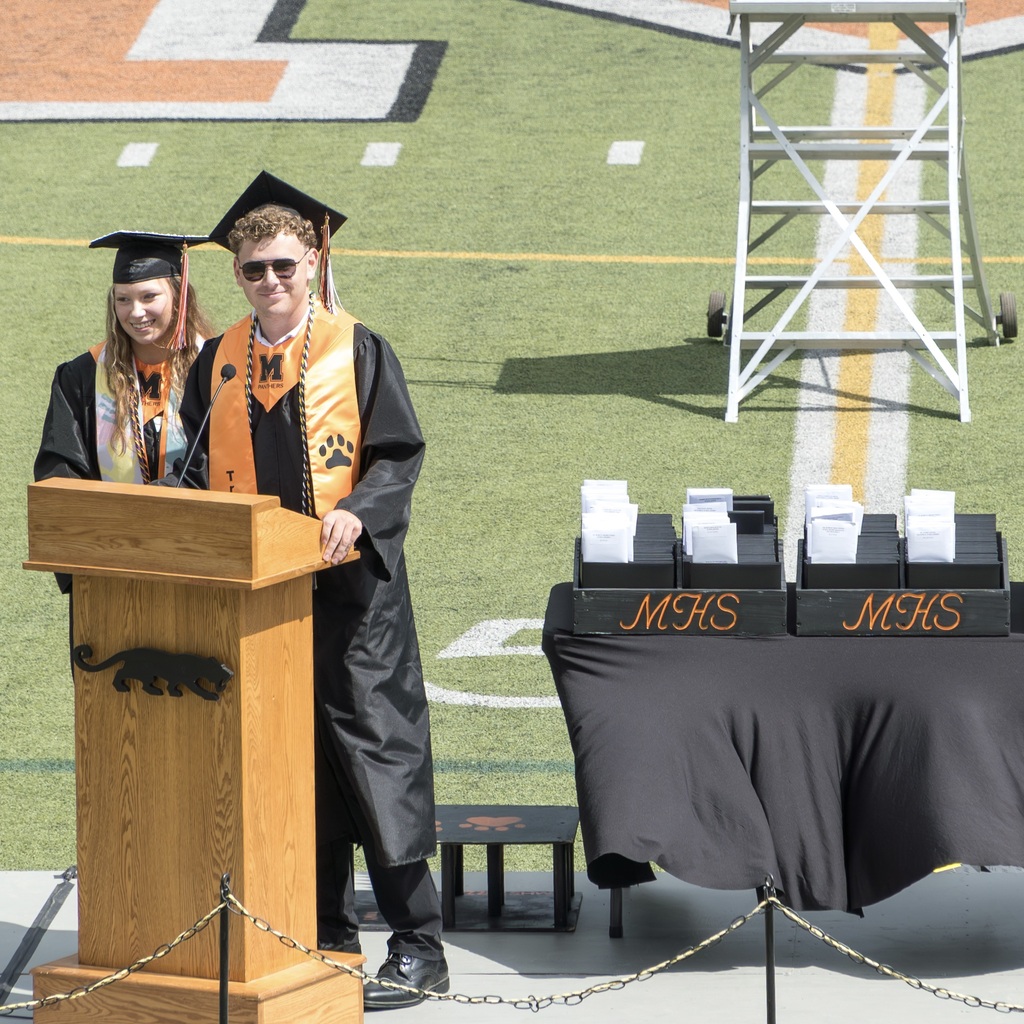 Two individuals in graduation robes stand at a podium on a football field with a covered table nearby.