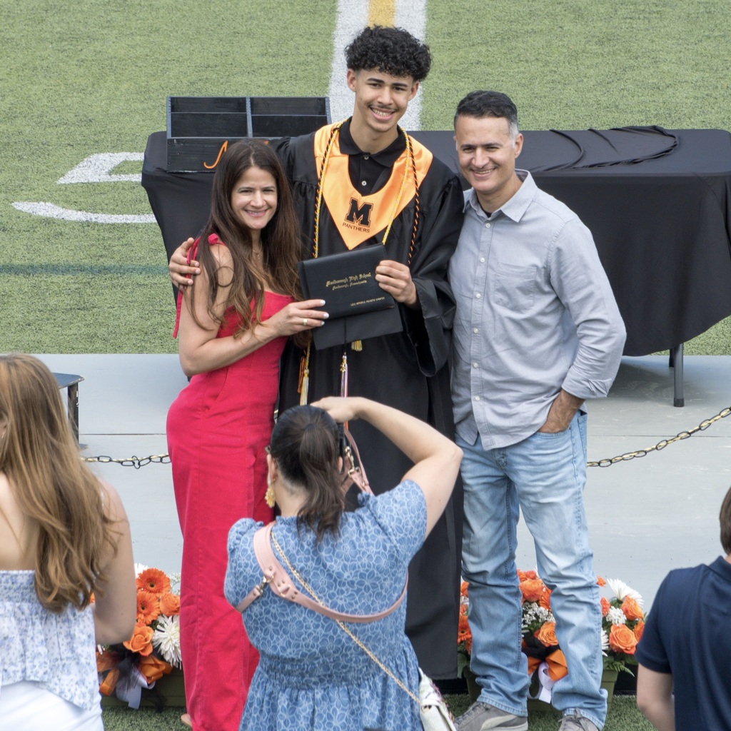 A girl in a red dress and a man in a blue button-down shirt and jeans pose for a group photo with an MHS graduate in a cap and gown standing in front of a platform with flowers.
