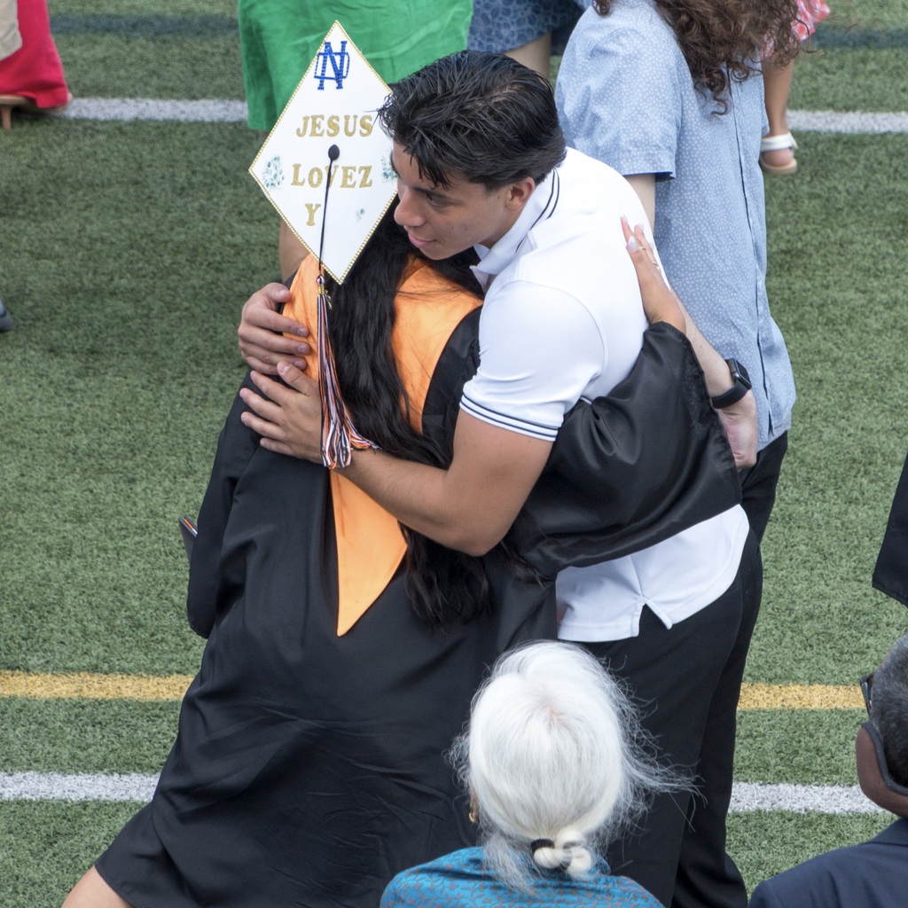 A man hugs a female MHS graduate in a cap and gown on a football field. They are surrounded by others.