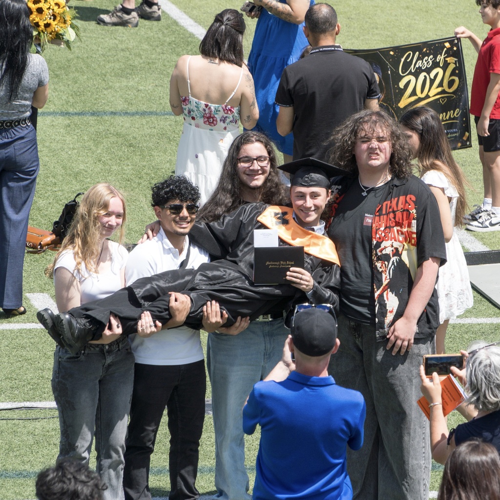 People in a field carry an MHS graduate across them for a group photo. A person is holding a sign that reads "Class of 2026" behind them.