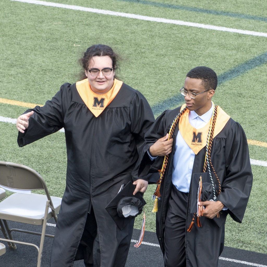 Two MHS students in black graduation gowns and caps walk together on a sports field.