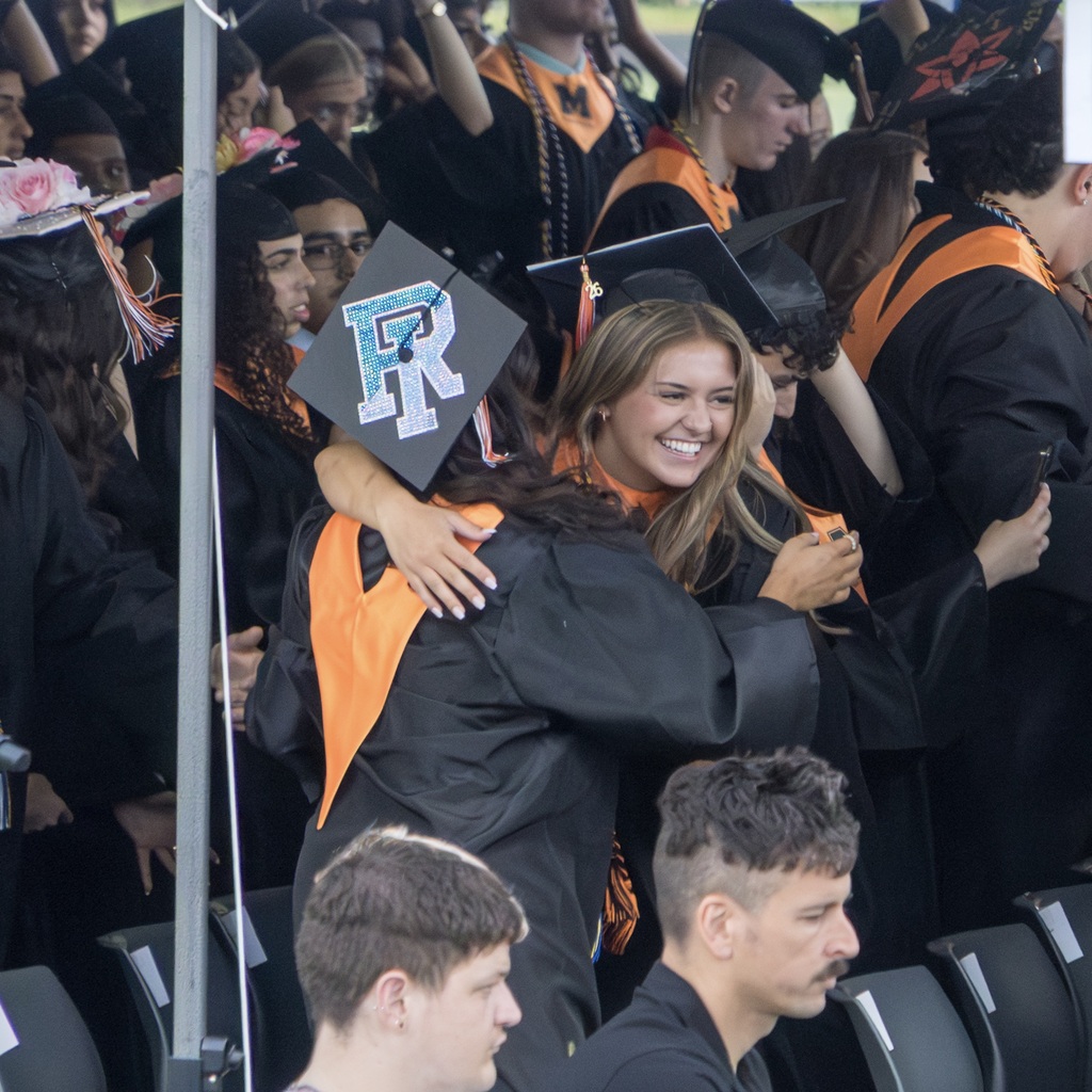 Group of MHS graduates in academic dress at an outdoor graduation ceremony. A smiling female graduate hugs another graduate.