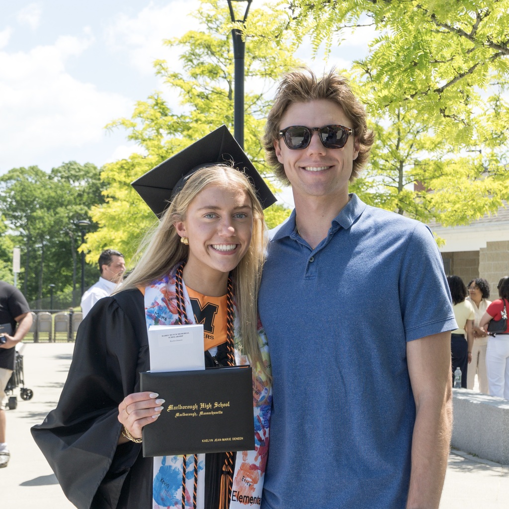 An MHS female graduate and a young man stand outside smiling for the camera; one in graduation attire holding a certificate, the other in a blue polo. Trees and buildings in the background.