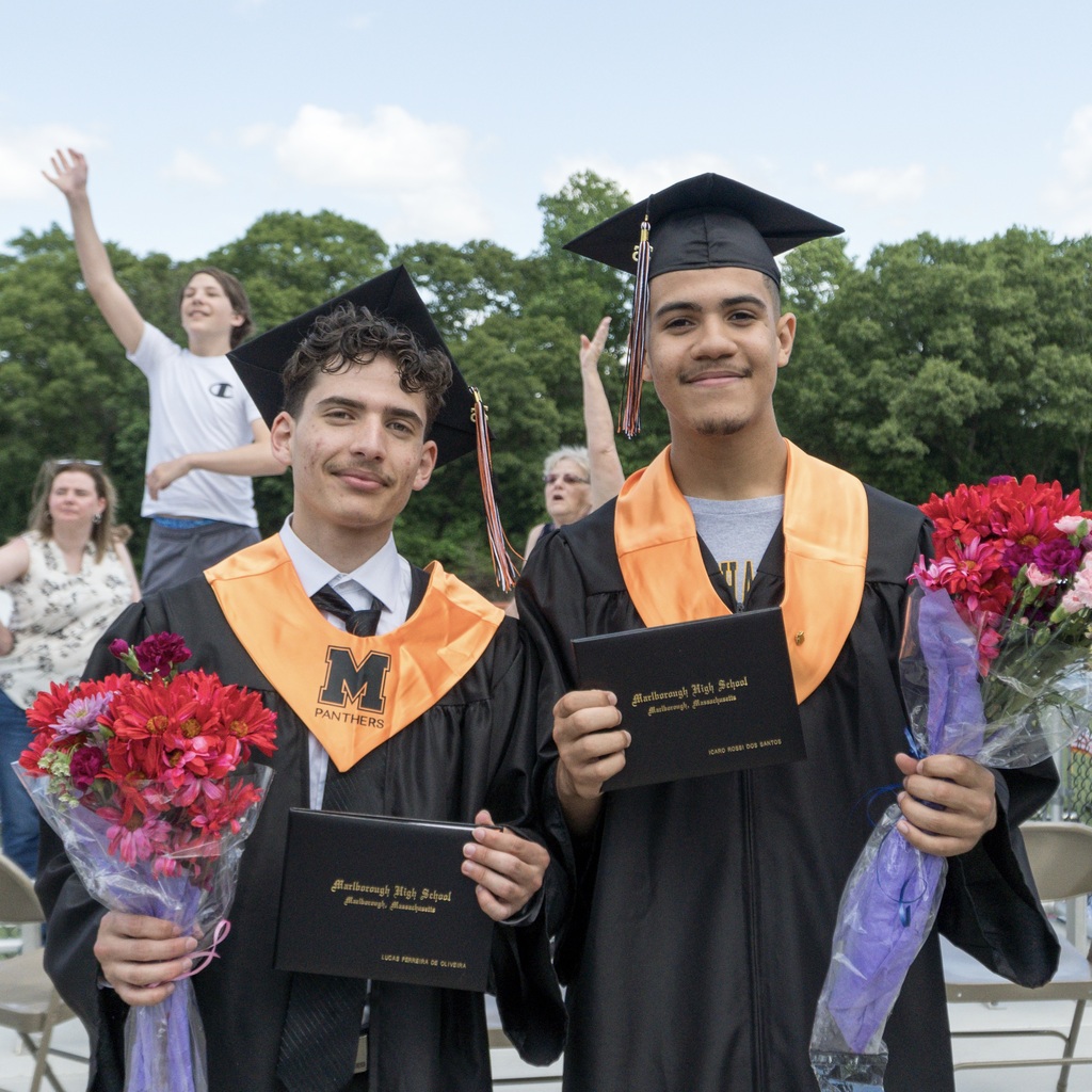 Two young men in graduation gowns and caps hold diplomas and bouquets. Trees and people in the background.