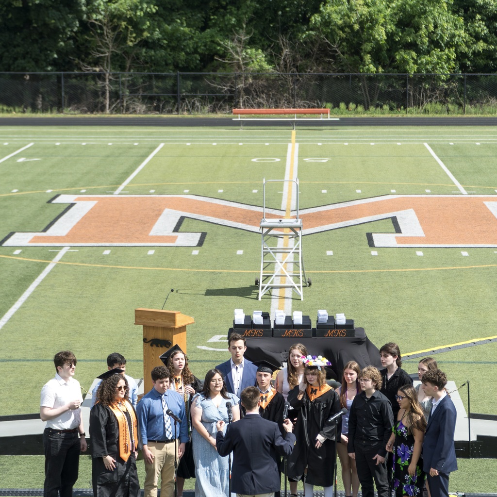 An MHS acapella group gathers on Noble Field to perform. A podium is behind them with a microphone.