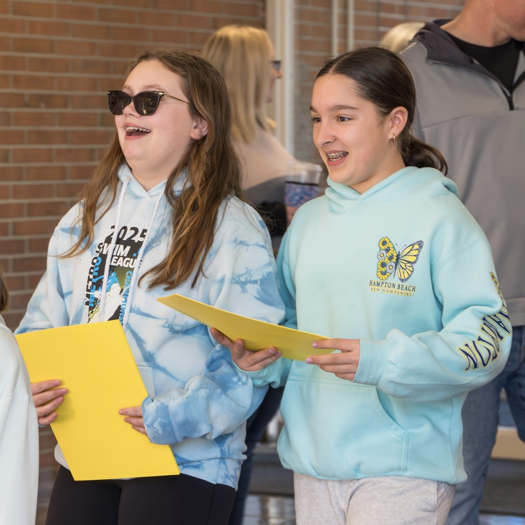 Two girls hold yellow folders, one in tie-dye and one in light blue, smiling and talking, while others walk in the blurred background.