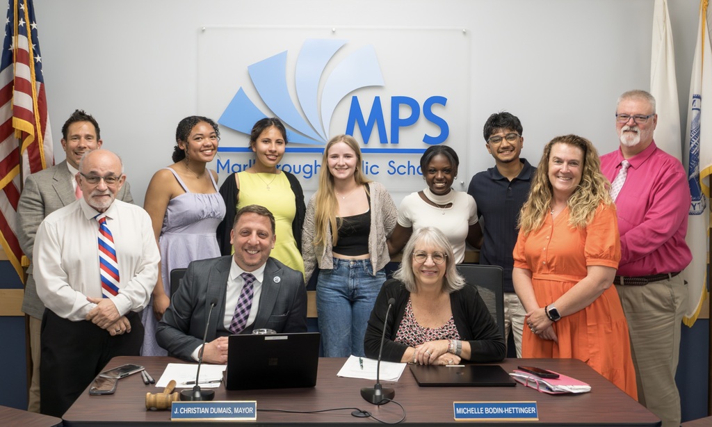 Six members of the Marlborough School Committee and the Student Advisory Council pose for a photo in front of a table with a laptop. Behind them, flags are displayed.