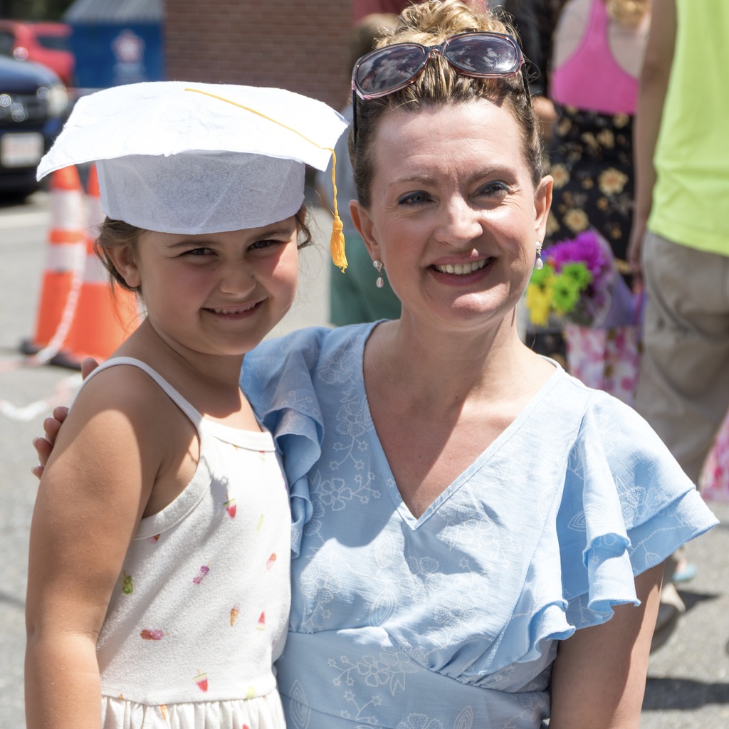 A woman and a girl stand close together, both smiling. The girl wears a graduation cap. Behind them, people and a traffic cone.