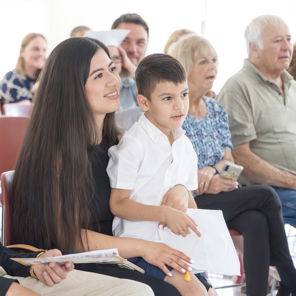 A boy holding a paper sits on his mother's lap. A group of adults and children sits in chairs, facing forward, with others seated next to and around them.