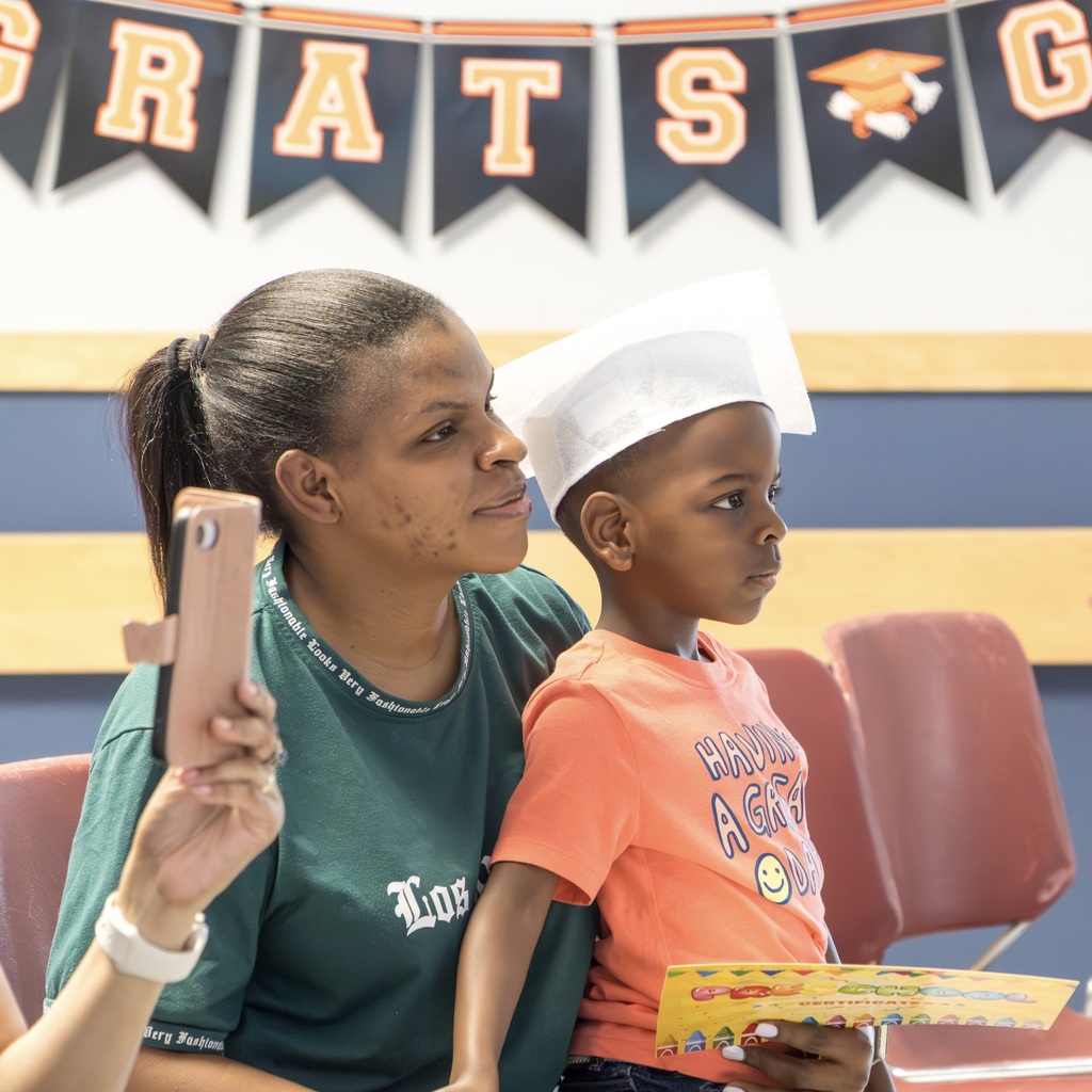 A woman and a child wearing a white graduation cap sit together, both holding items. Behind them is a banner reading "CONGRATS."