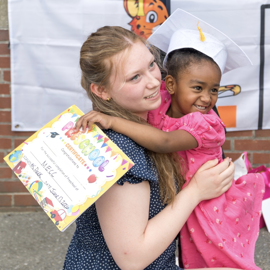 A young girl in a graduation cap and pink dress holds a certificate while hugging a female teacher in a navy blue polka-dot dress.
