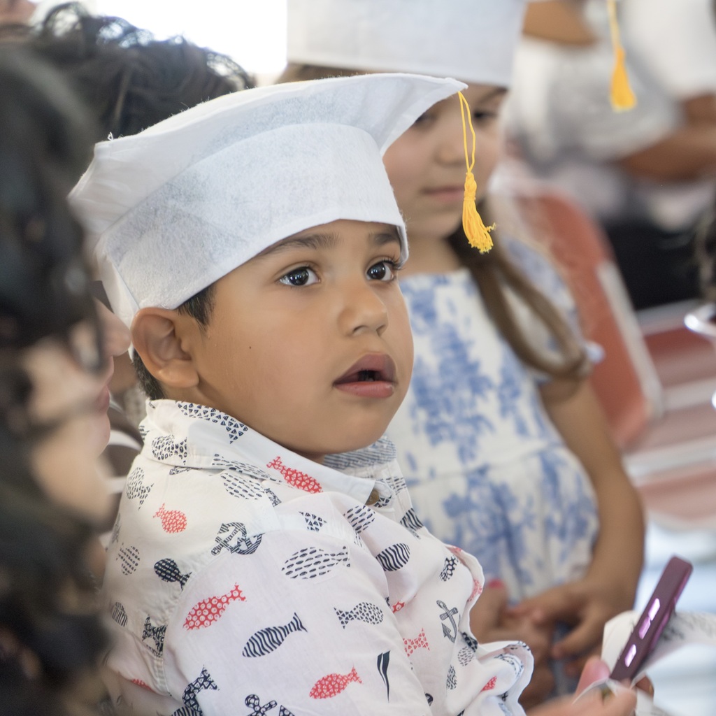 A child in a graduation hat and fish print shirt, looking up, sitting next to another child in a white graduation gown.