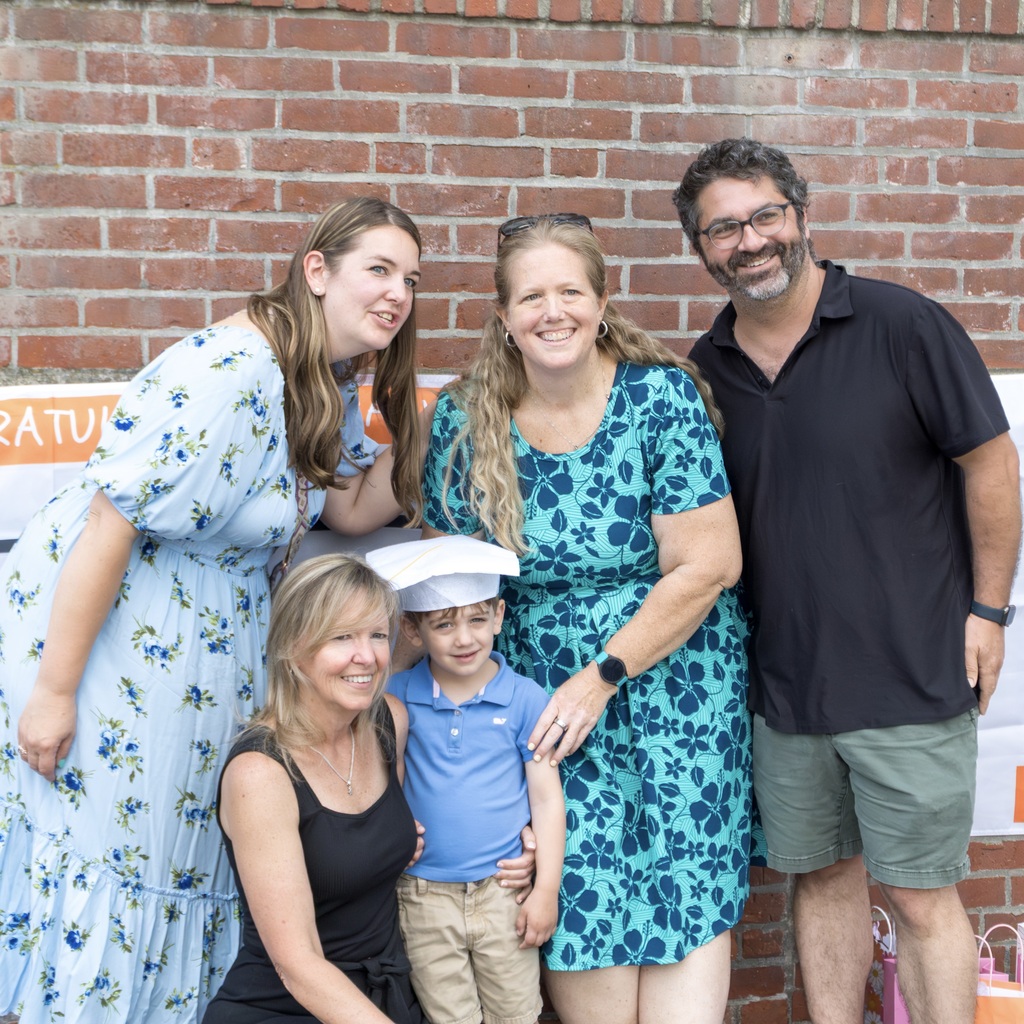 Four adults and a young boy wearing a white graduation cap pose for a photograph in front of a brick wall.
