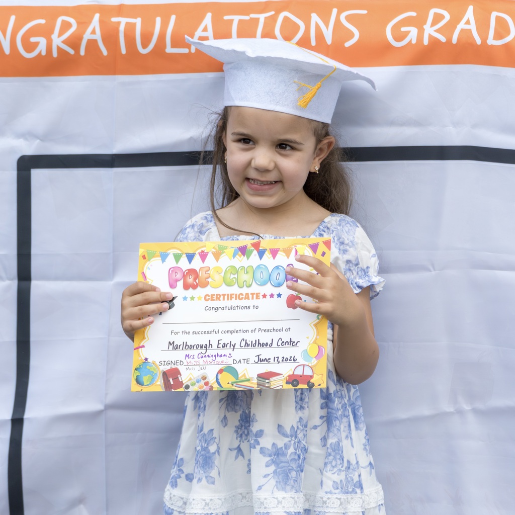 A young girl wearing a graduation cap smiles as she holds a certificate. Background shows a banner reading "Congratulations Grad".