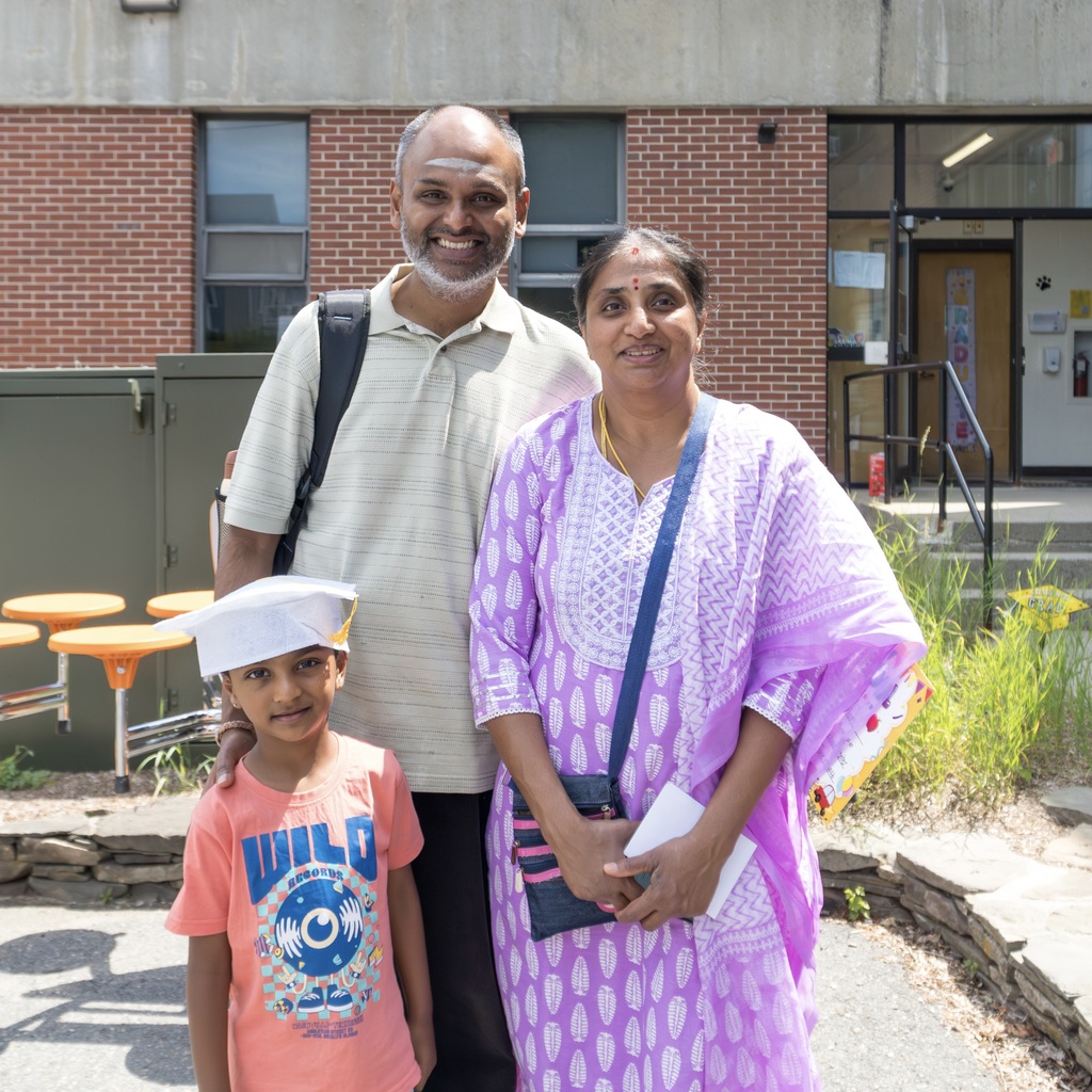 A man and a woman stand outside with a child wearing a graduation cap. They smile for the camera.
