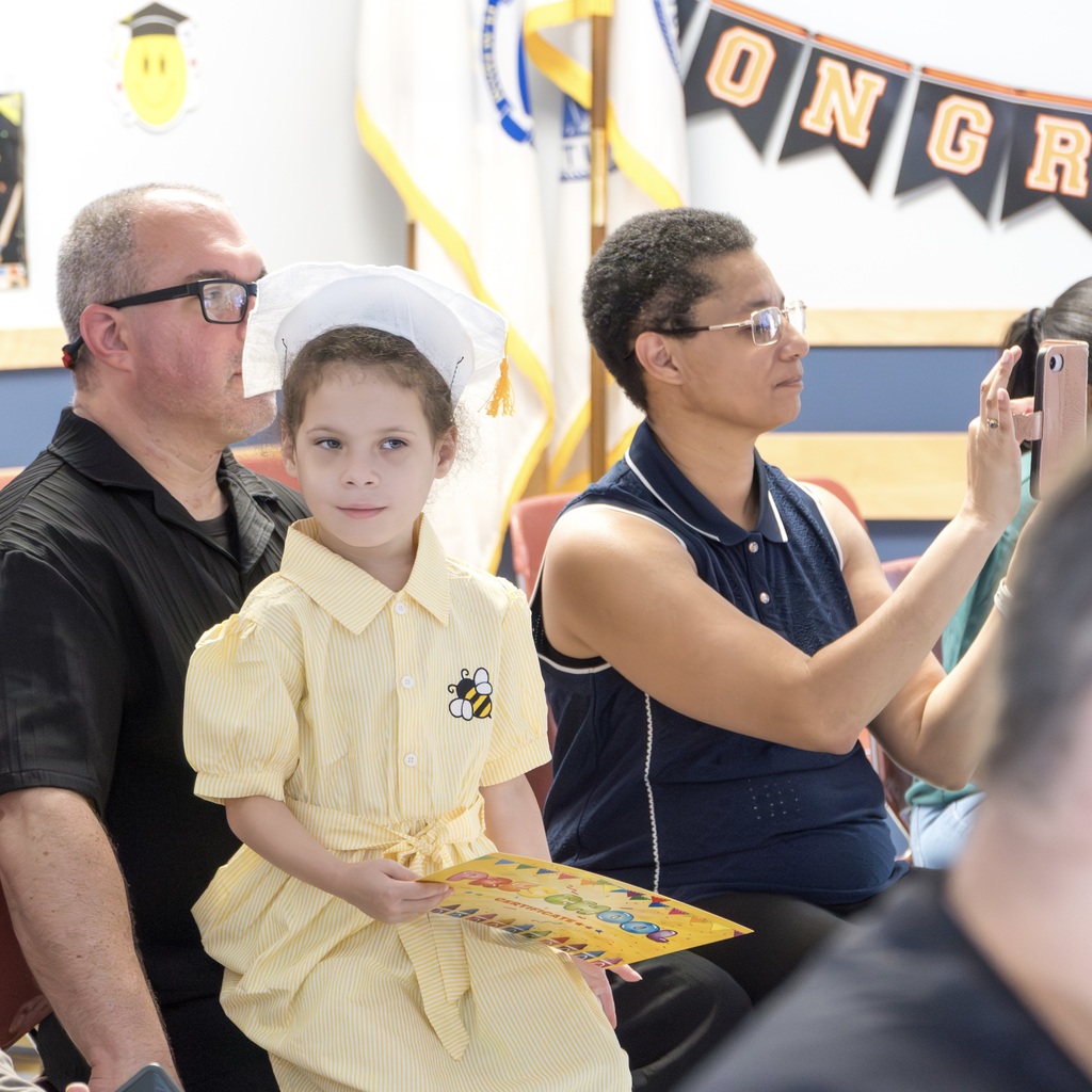 Three people sit together in a room; a young girl in a yellow dress holds a certificate. A flag and banner are in the background.