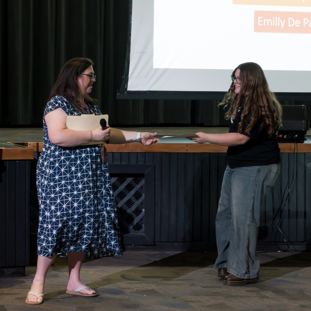 A Whiticomb teacher in a dress stands and presents an award to a female student in jeans and a shirt.