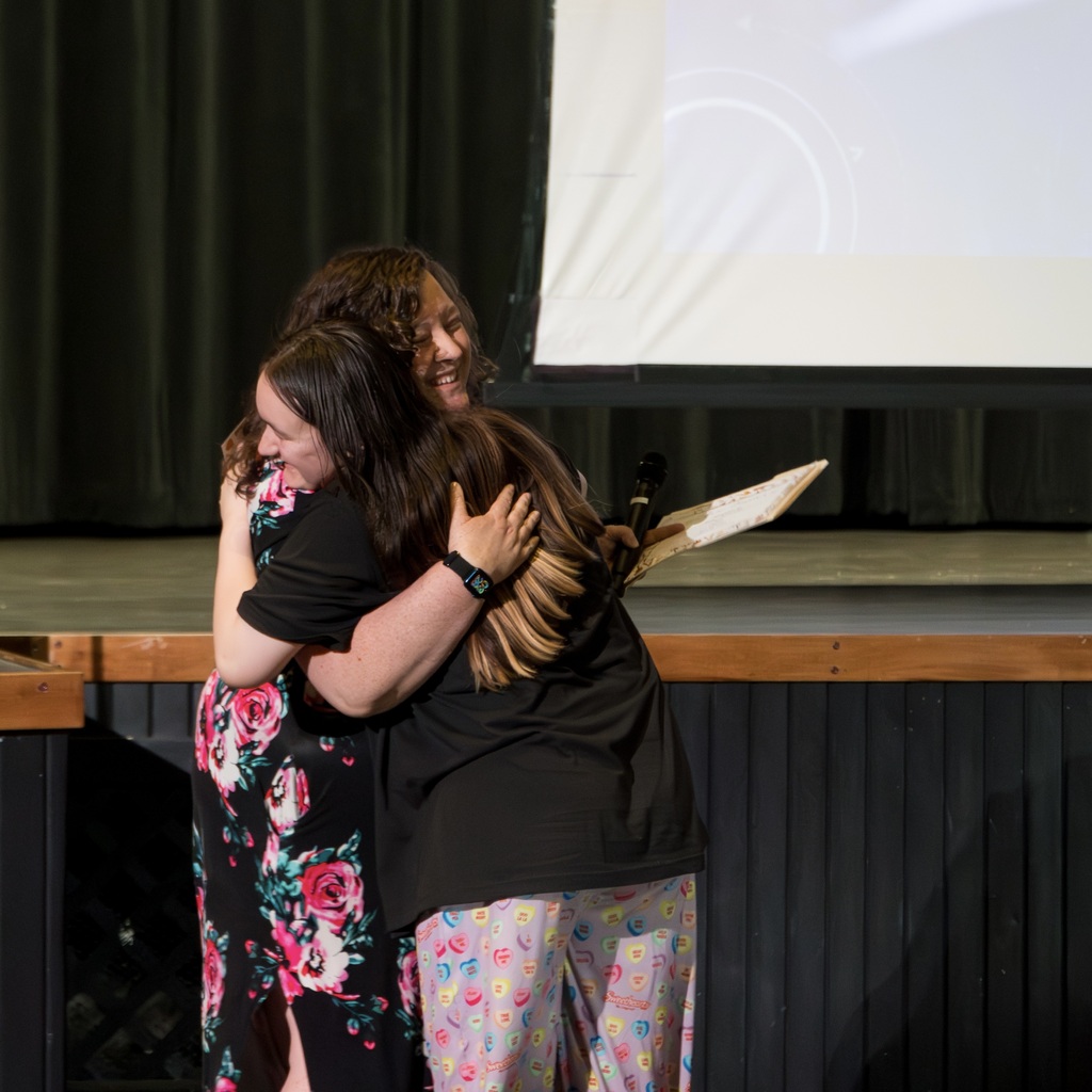 A teacher and a student embrace on a stage; one wears a floral dress, the other a black shirt. A large screen is behind them.