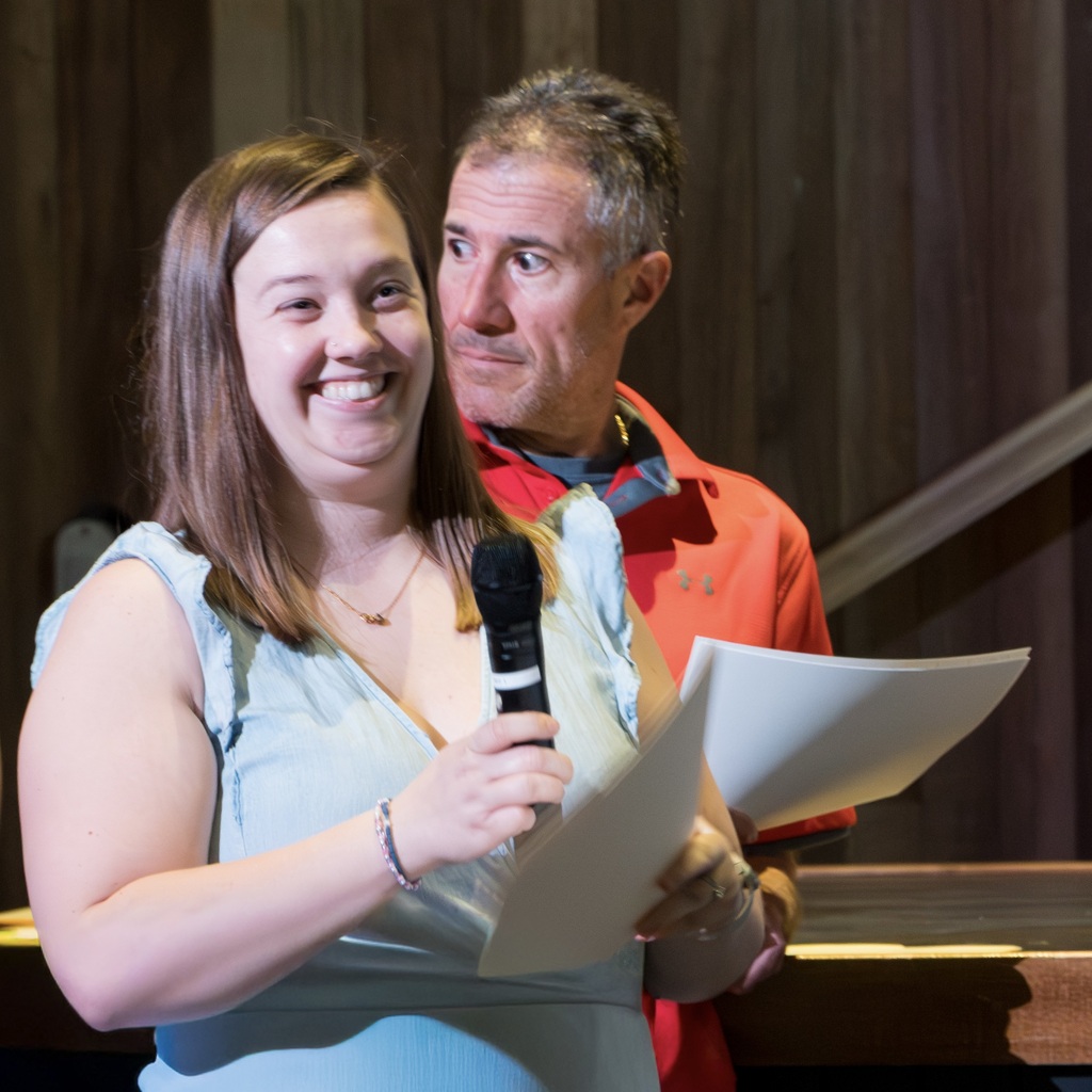 A Whitcomb teacher holding a microphone and papers, smiling. A man behind her holds papers as they both stand.