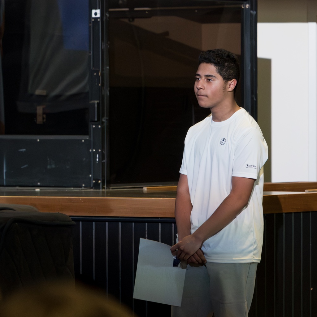 A young man in a white shirt stands on a stage, holding a paper. Behind him, a stage.