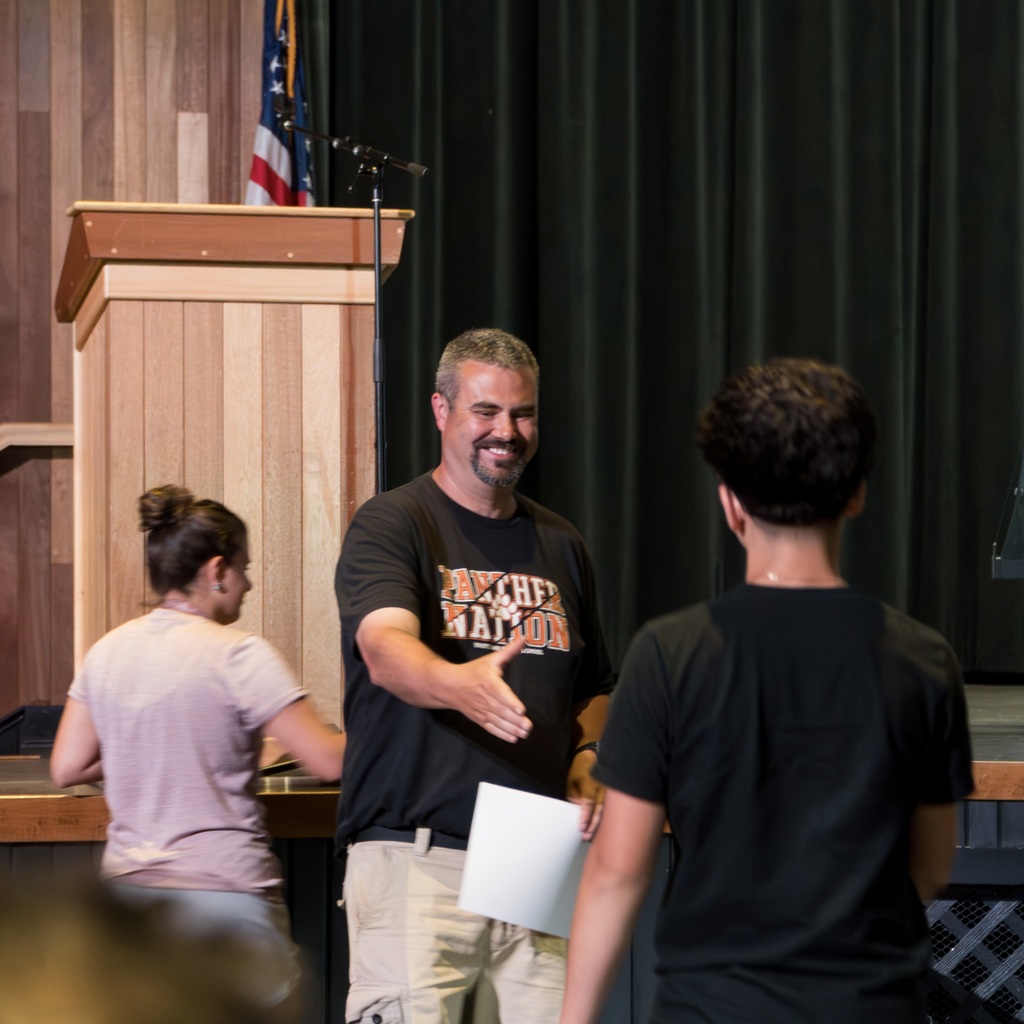 A smiling male Whitcomb teacher extends his hand to shake a student's hand as the student walks toward him to receive a certificate. A female teacher stands behind the male teacher.