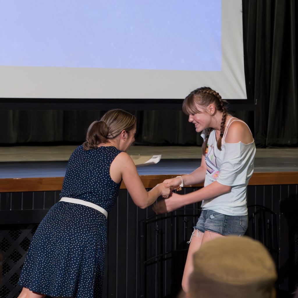 A Whitcomb female teacher shakes hands with a female student. The teacher wears a blue dress; the other, a light blue shirt. Black curtains and a projector screen are in the background.
