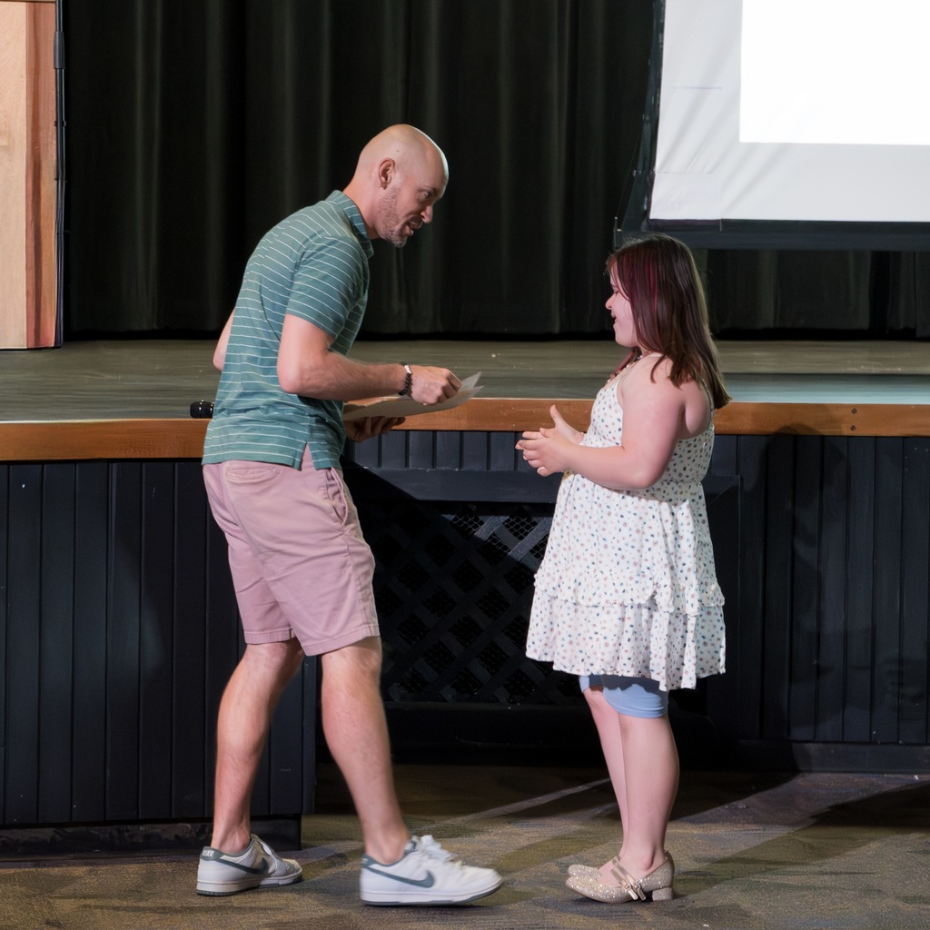 A male teacher and a young female student are standing; the man appears to be presenting the woman with a certificate.