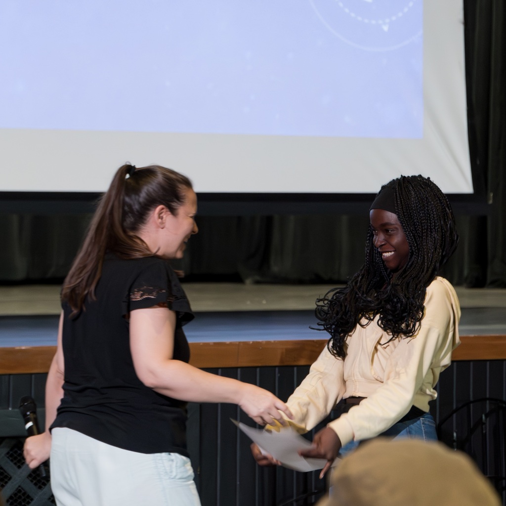 A Whitcomb teacher and a student stand in a room, both smiling, one holding a paper. A projector screen behind them shows a blue screen.