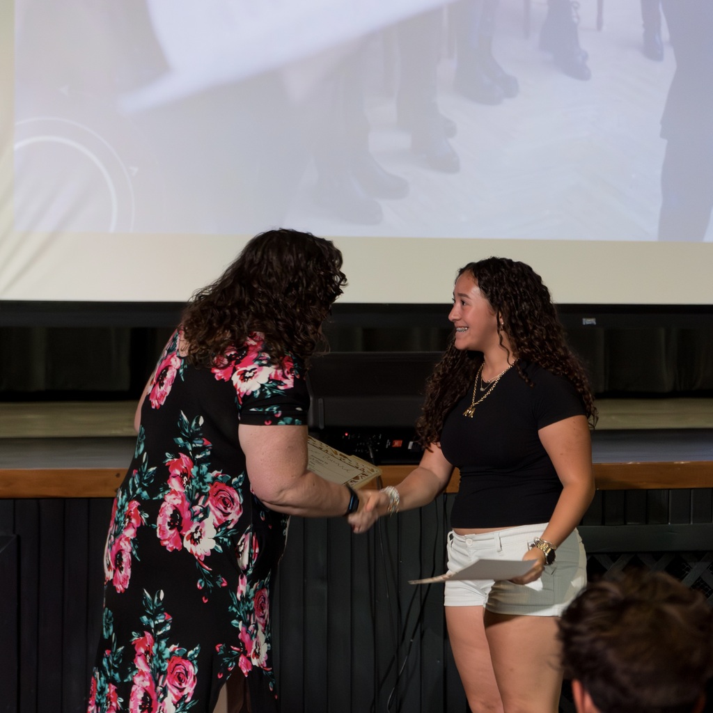A teacher n a floral dress and a student in a black shirt and white shorts stand shaking hands. A projection screen behind them shows blurred images.