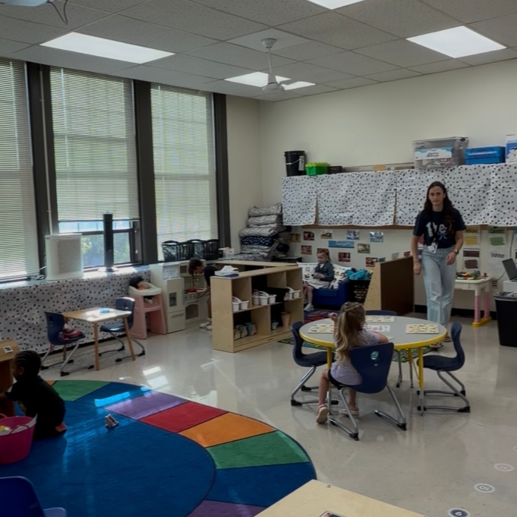 A room with white walls and large windows. A woman stands near a table, and children sit at tables and play on a colorful rug.