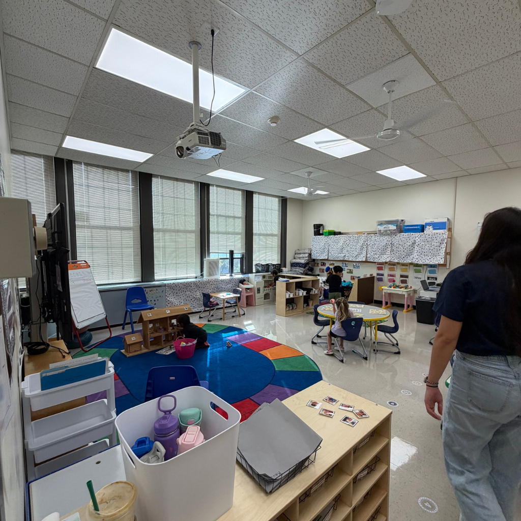 Bright classroom with white walls and large windows. A person stands near a table with toys and a rug.