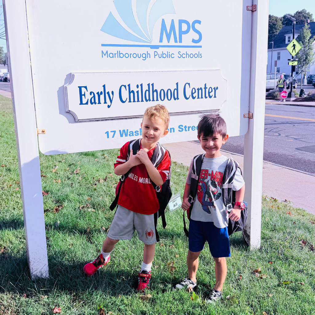 Two young boys stand in front of a sign that reads "Early Childhood Center" with a road behind them.