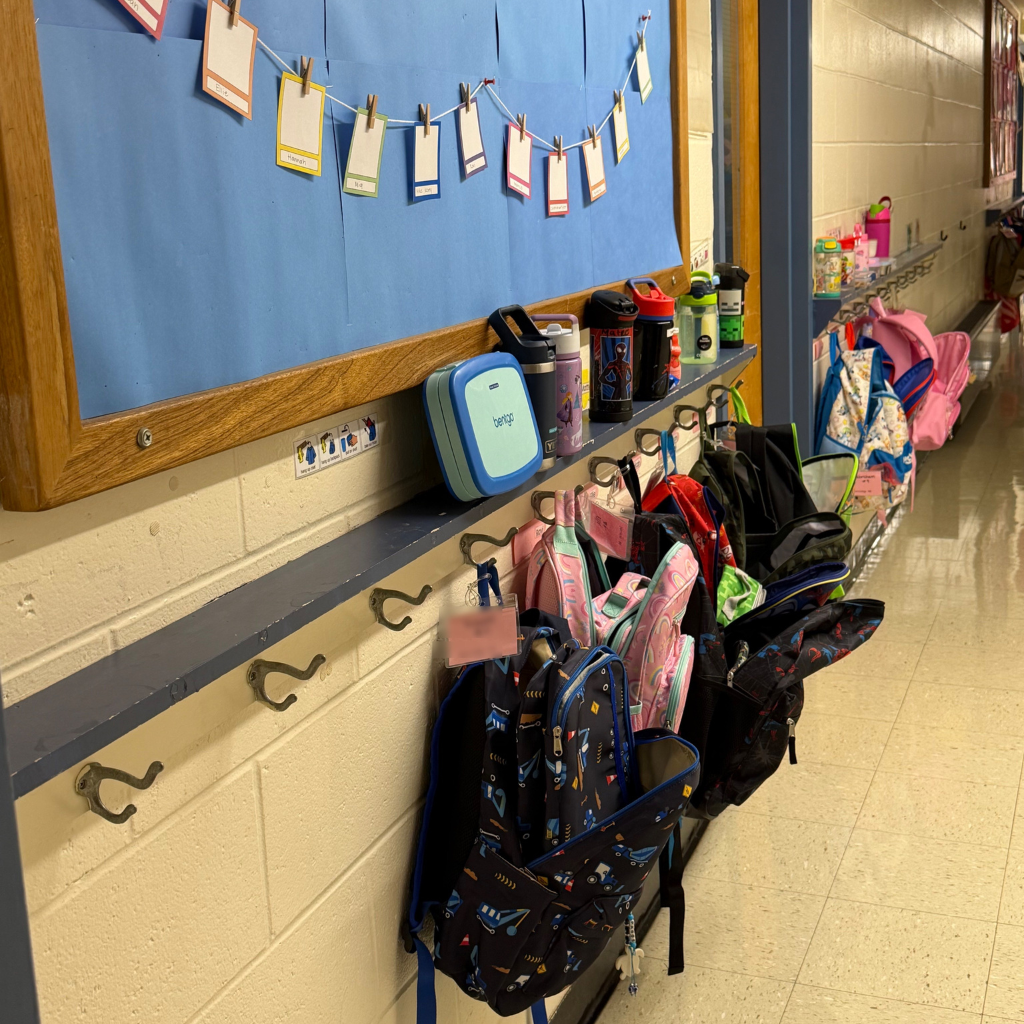 Hallway with a bulletin board and name tags, hooks with backpacks, a lunchbox, and bottles.