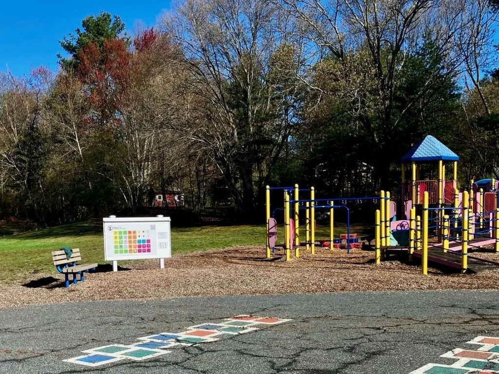 A playground with climbing structures, slides, and a hopscotch pattern on the ground. A sign is beside the bench. Trees and grass surround the area.