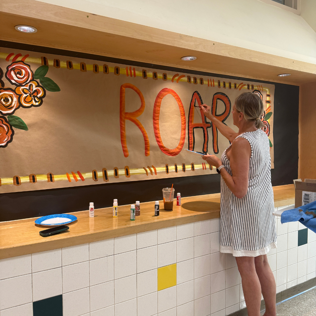 A woman stands at a counter, painting the word "ROAR" on a banner featuring flowers.