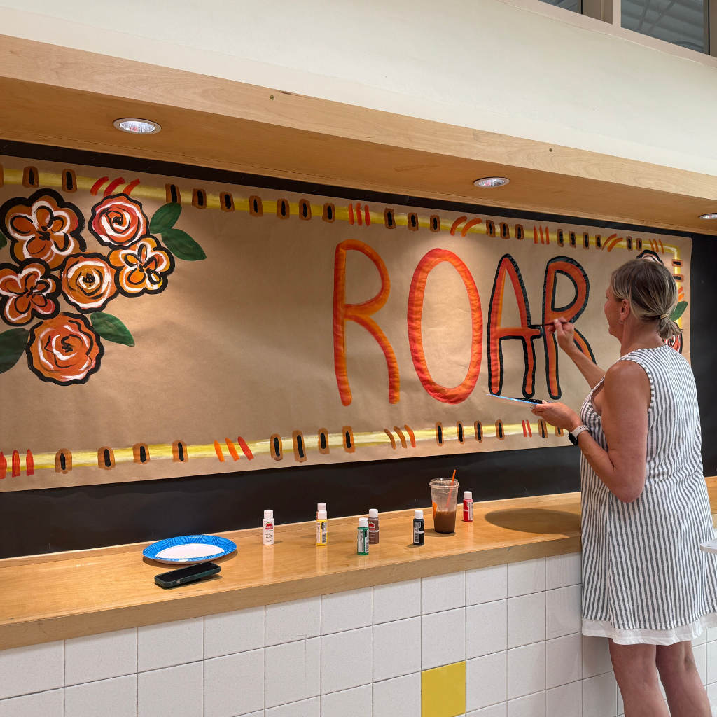 A woman painting a banner with "ROAR" and flowers on a wall. Tiled wall in background.