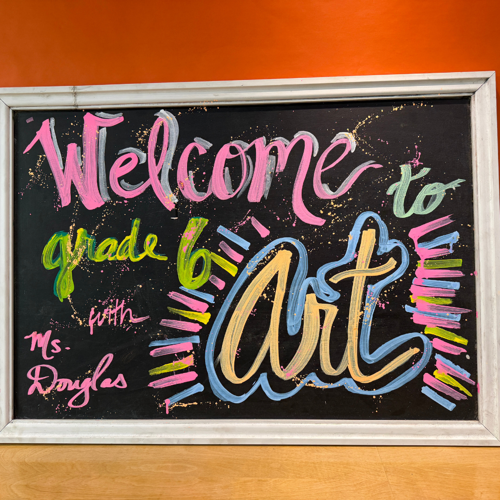 A blackboard with multicolored chalk. The words "Welcome to grade 6 Art" are written. Below it, "with Ms. Douglas" is visible.