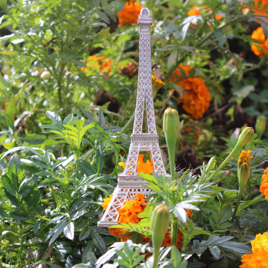 A small, white replica of the Eiffel Tower stands among green foliage and marigold flowers, with orange petals visible.