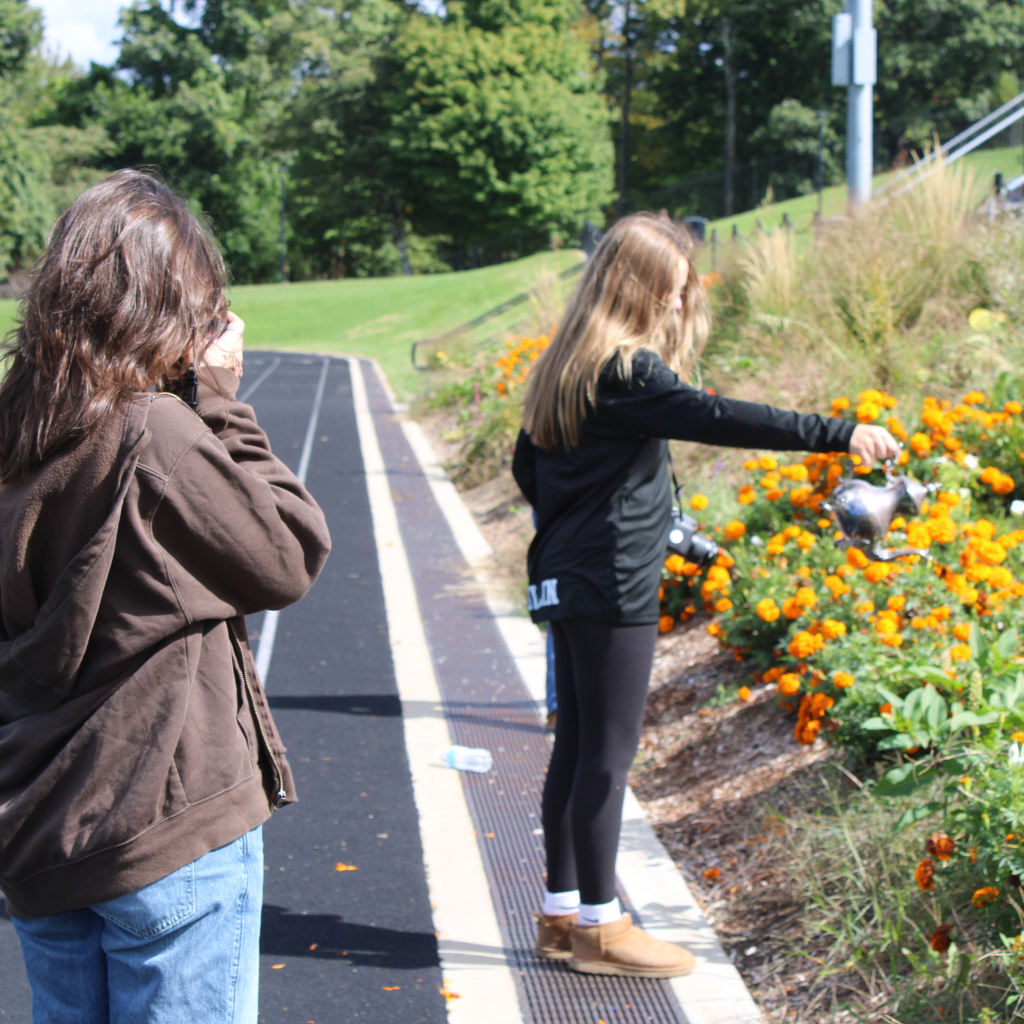 A woman stands on a track while another waters orange flowers on a sloped grassy area.
