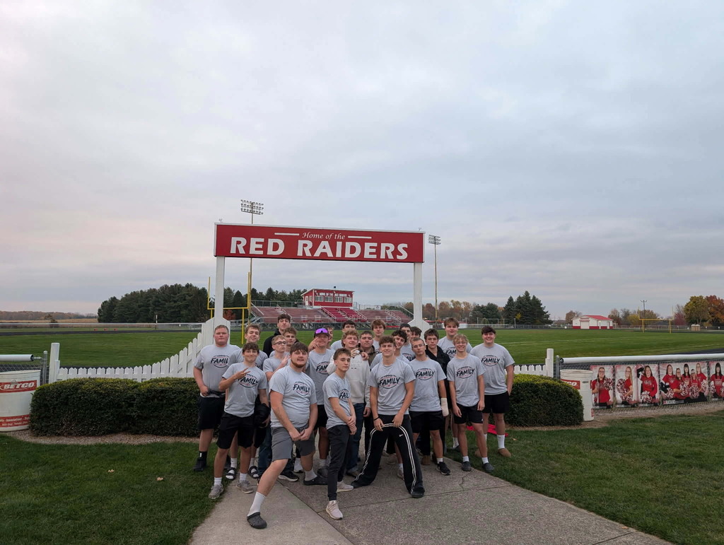 The Red Raiders football team poses for a group photo in front of a red sign that reads “Home of the Red Raiders” at their football field. The players wear matching gray shirts with “Family” printed on the front.