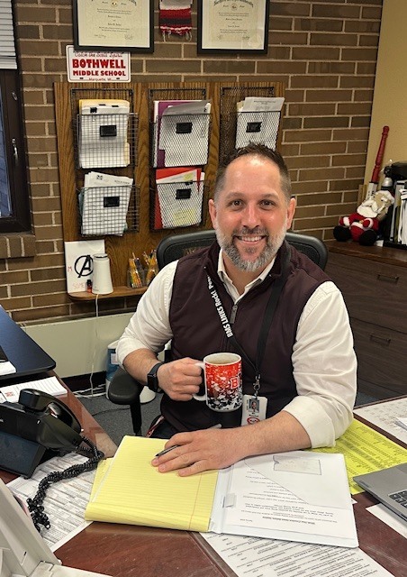 Photo of Mr. Reichel at his desk holding a BMS coffee mug , showing he is ready to start the day!