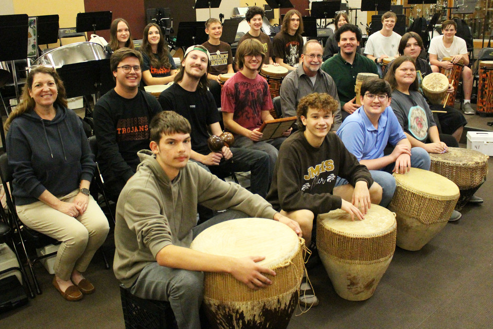 Large group of people sitting and standing in a music room with drums, focused on playing hand drums and percussion instruments together.