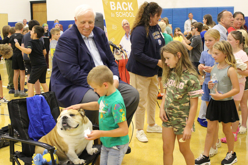 A group of people, including children, gathered around a large dog in a school gymnasium. One man in a suit appears to be speaking to the group.