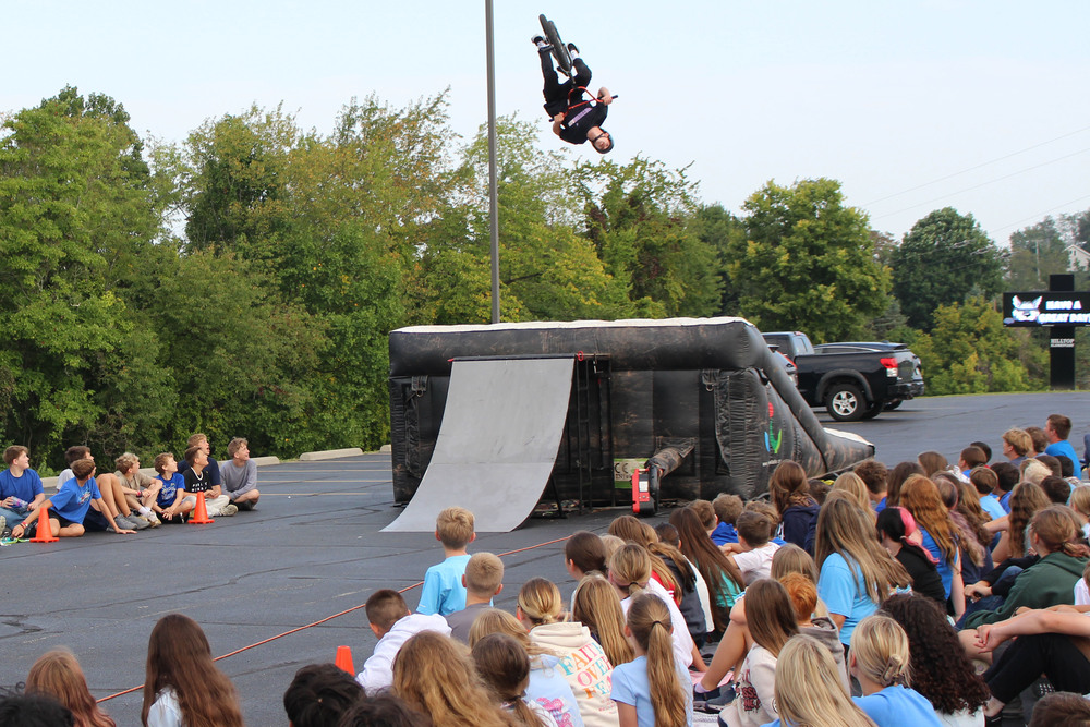 BMX biker performing trick in front of crowd of spectators seated on pavement near vehicles.