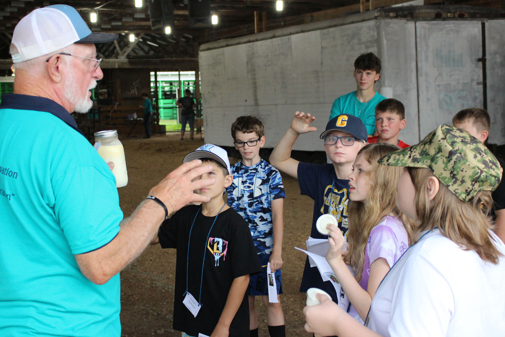 A group of young people listening attentively to an older man wearing a cap and casual clothing in a large indoor space.