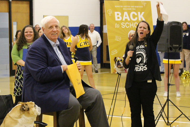 A senior man in a suit sitting in a chair, surrounded by people, including a woman raising a sign that says "Back to School".